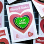 A green heart-shaped enamel pin with the words 'LAST WORD' in white lettering, displayed on a white backing card with red and pink heart graphics. The card is placed on top of other similar cards on a black grid cutting mat background.